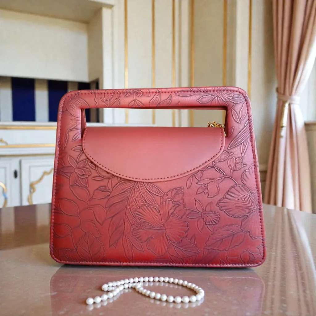 Brick red embossed floral mini crossbody bag on table showing top handle cutout gold chain strap and textured floral pattern