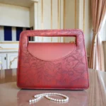 Brick red embossed floral mini crossbody bag on table showing top handle cutout gold chain strap and textured floral pattern