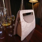 Side angled view of soft pink embossed floral mini carry crossbody handbag with chain strap placed on wooden dining table with glassware