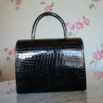 Back view of glossy black croc textured top handle handbag on a marble surface with floral wallpaper in the background