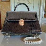 Glossy black croc top handle handbag with gold buckle displayed on glass surface alongside pearl bracele