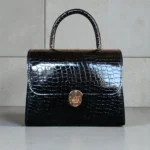 Glossy black croc textured top handle handbag with gold turn-lock buckle displayed on a light stone background