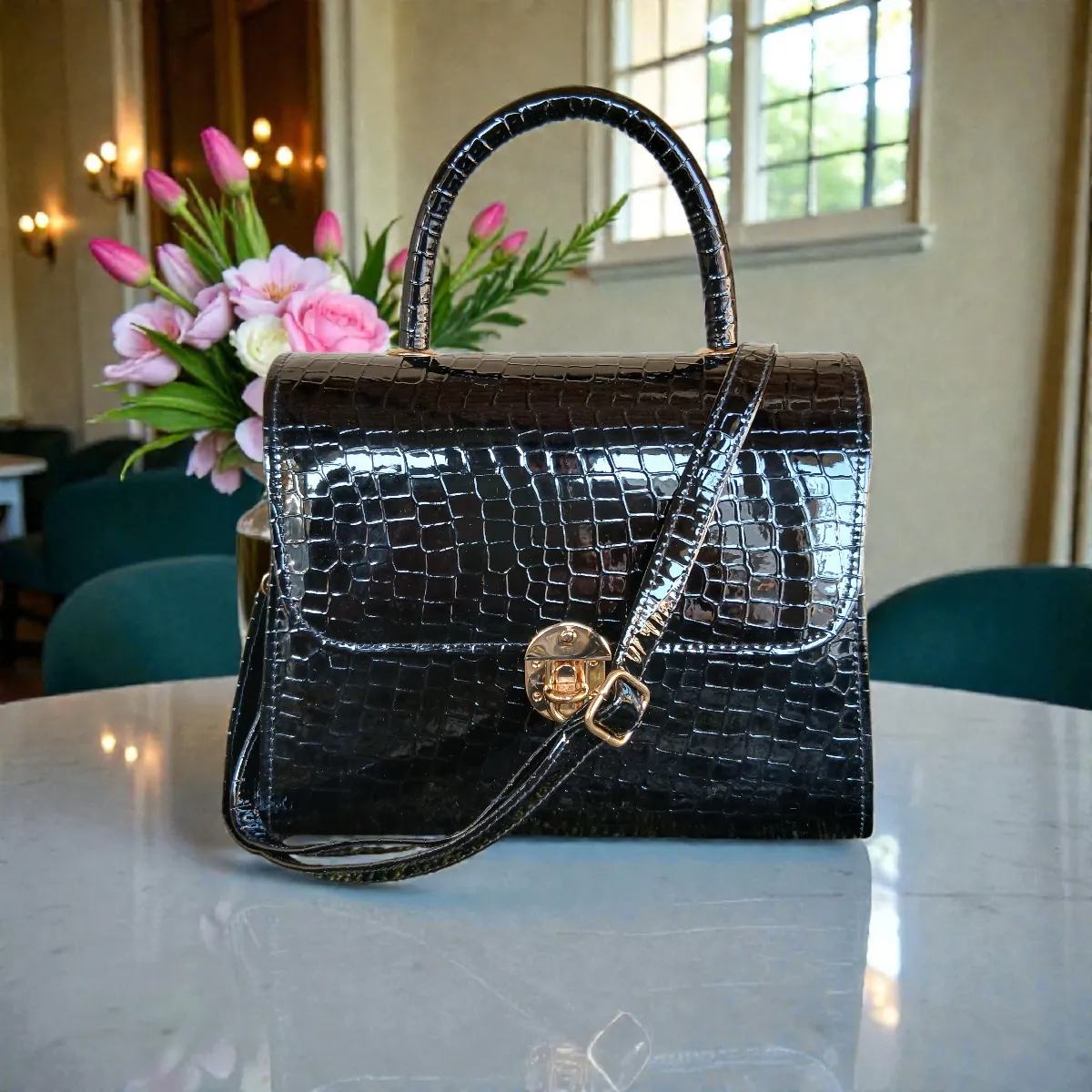 Glossy black croc handbag with top handle and adjustable strap placed on a table with white flower vase behind