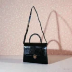 Glossy black croc top handle handbag hanging by adjustable shoulder strap against patterned pink background