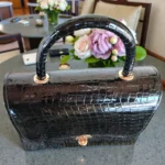 Top close-up of glossy black croc handbag showing handle attachments and gold metal hardware details