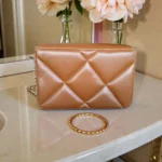 Tan brown quilted crossbody bag with pearl chain detail placed on a marble vanity near flowers.