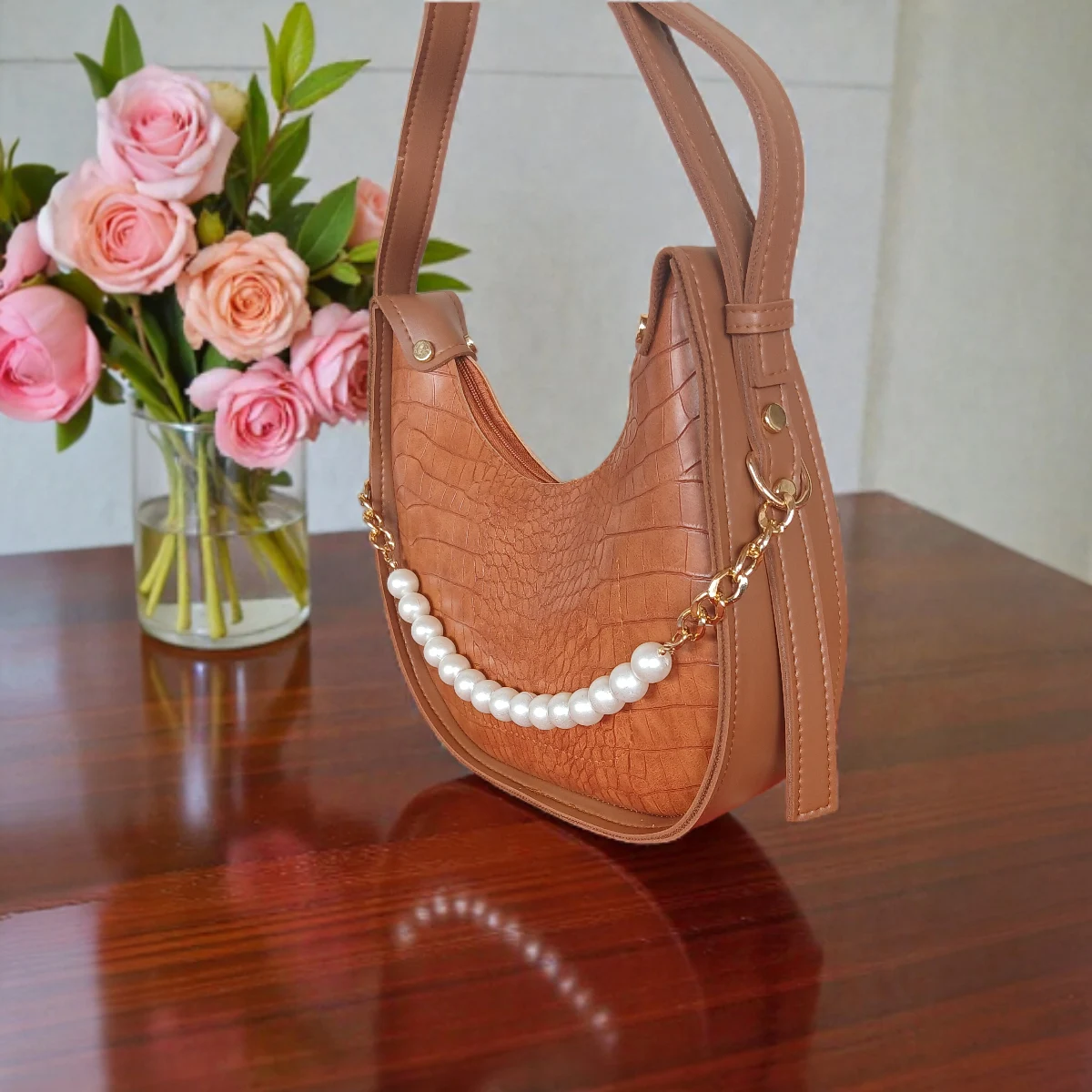 Textured tan brown crossbody bag with pearl chain displayed on wooden table, angled view