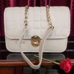 Front display of ivory white embossed grid leather flap crossbody bag with gold chain strap placed on wooden table with rose