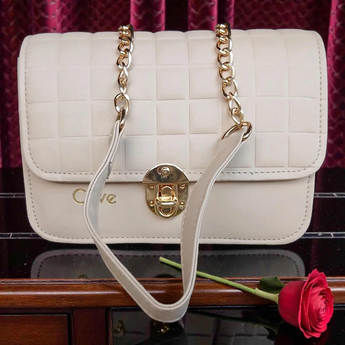 Front display of ivory white embossed grid leather flap crossbody bag with gold chain strap placed on wooden table with rose