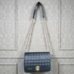 Jet Black embossed grid leather flap chain crossbody bag with gold chain strap hanging against textured wall background