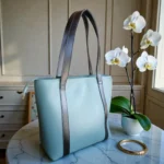 Minimal dusty blue tote handbag with black straps side angle view placed near orchid flowers